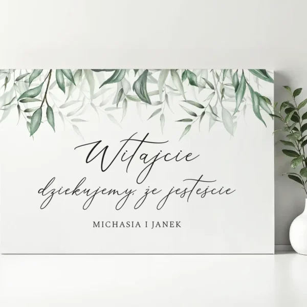 Elegancka tablica powitalna z napisem "Witajcie, dziękujemy, że jesteście" ozdobiona motywem zielonych gałązek, ustawiona na tle jasnej ściany z dekoracyjną rośliną w białej doniczce.