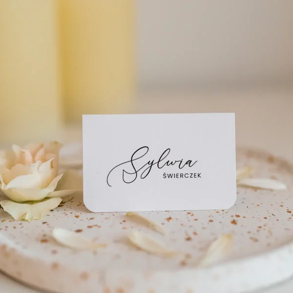 Winietki na roczek z eleganckim, minimalistycznym designem, ułożone na ozdobnym talerzu w otoczeniu kwiatów i płatków róż.