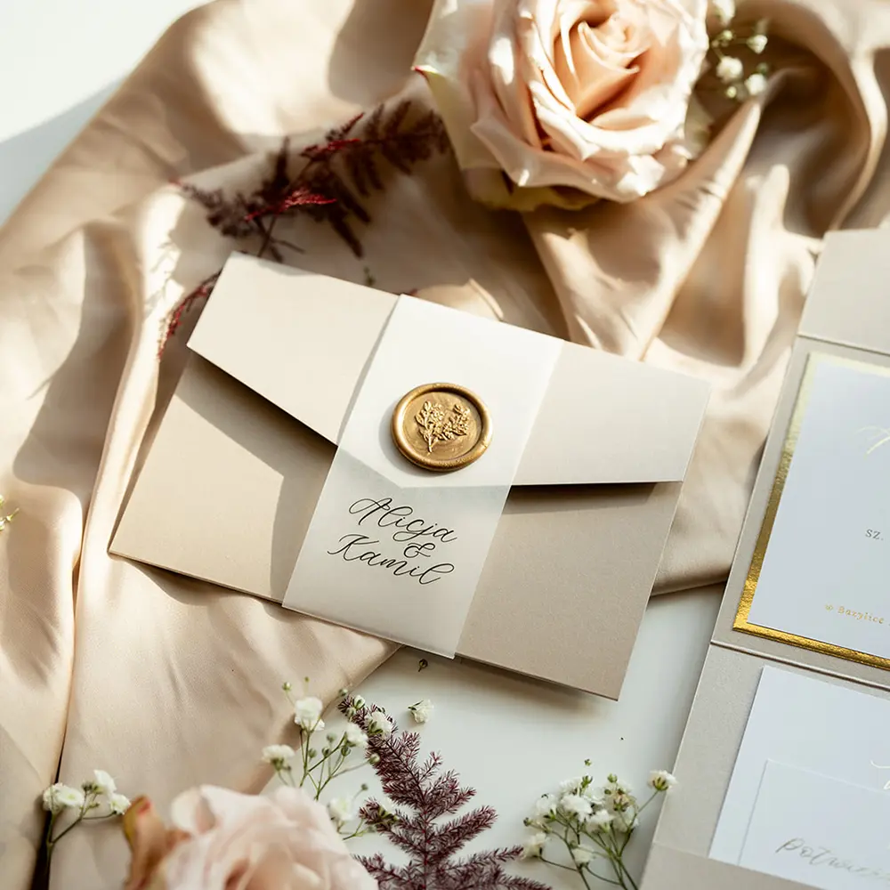 Eleganckie zaproszenie ślubne w stylu glamour, w beżowej tonacji z dekoracyjną opaską i woskową pieczęcią w złotym odcieniu. Zaproszenie ułożone na luksusowej, satynowej tkaninie, w otoczeniu subtelnych kwiatów, podkreślających romantyczny charakter papeterii.