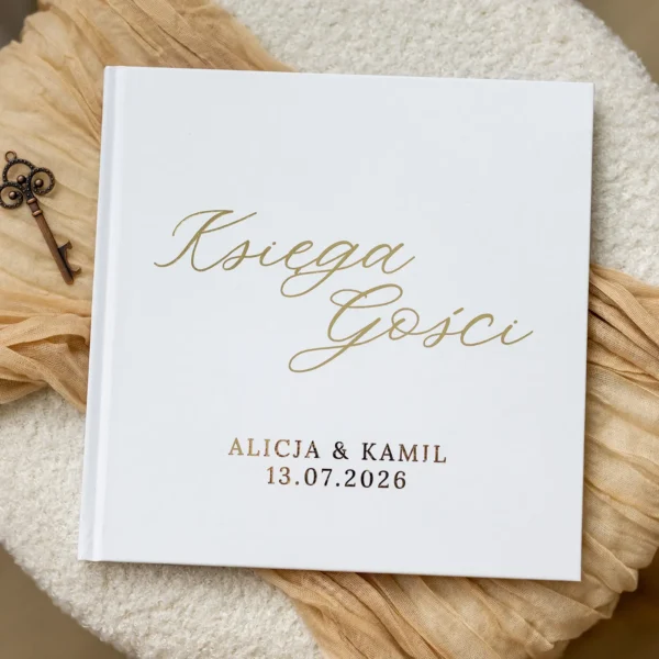 Elegancka biała księga gości ślubnych z napisem w złotym kolorze, ułożona na kremowej tkaninie obok dekoracyjnego klucza.