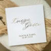 Biała księga gości weselnych z personalizowanym złotym napisem na okładce, ułożona na kremowej tkaninie i ozdobiona dekoracyjnym kluczykiem.