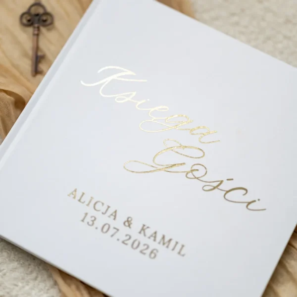 Zbliżenie na białą księgę gości z połyskującym złotym napisem i datą ślubu, na tle jasnej tkaniny i ozdobnego klucza.