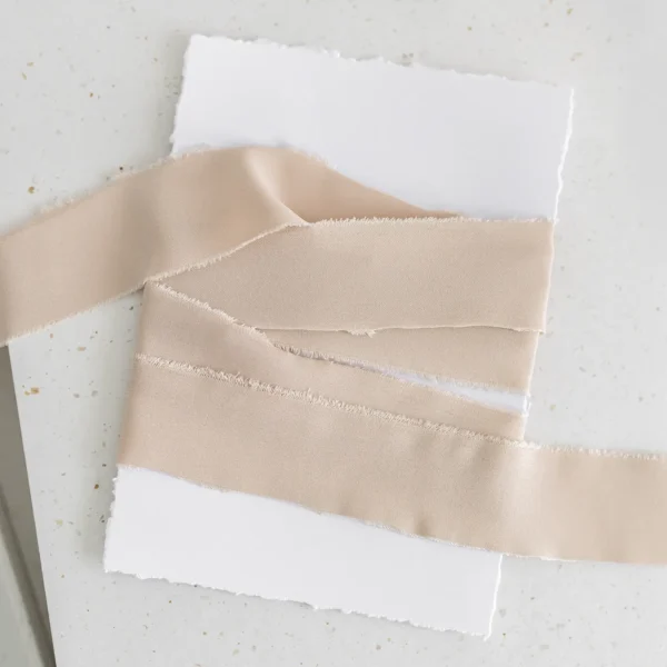 Beżowa wstążka satynowa owinięta wokół białej kartki o postrzępionych brzegach.