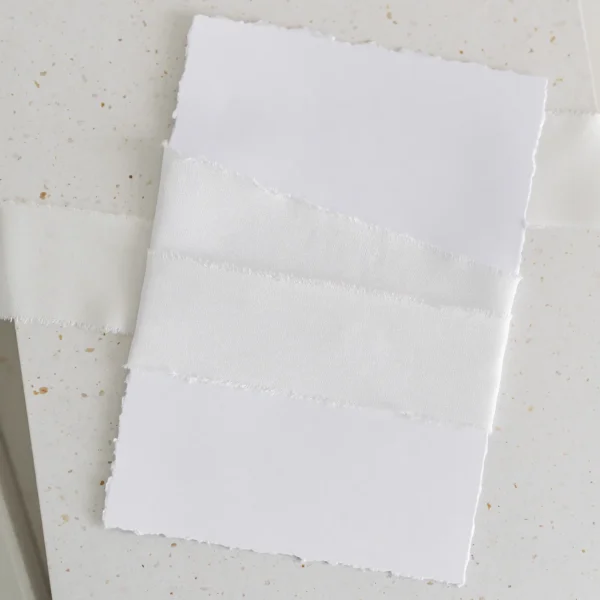 Biała wstążka satynowa owinięta wokół białej kartki o postrzępionych brzegach.