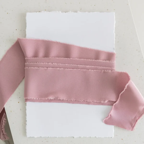 Ciemnoróżowa wstążka satynowa owinięta wokół białej kartki o postrzępionych brzegach.