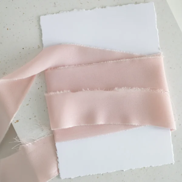 Jasnoróżowa wstążka satynowa owinięta wokół białej kartki o postrzępionych brzegach.