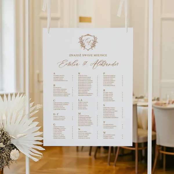 Plan stołów z alfabetycznym układem nazwisk i złotym monogramem, zawieszony na białej ramie.