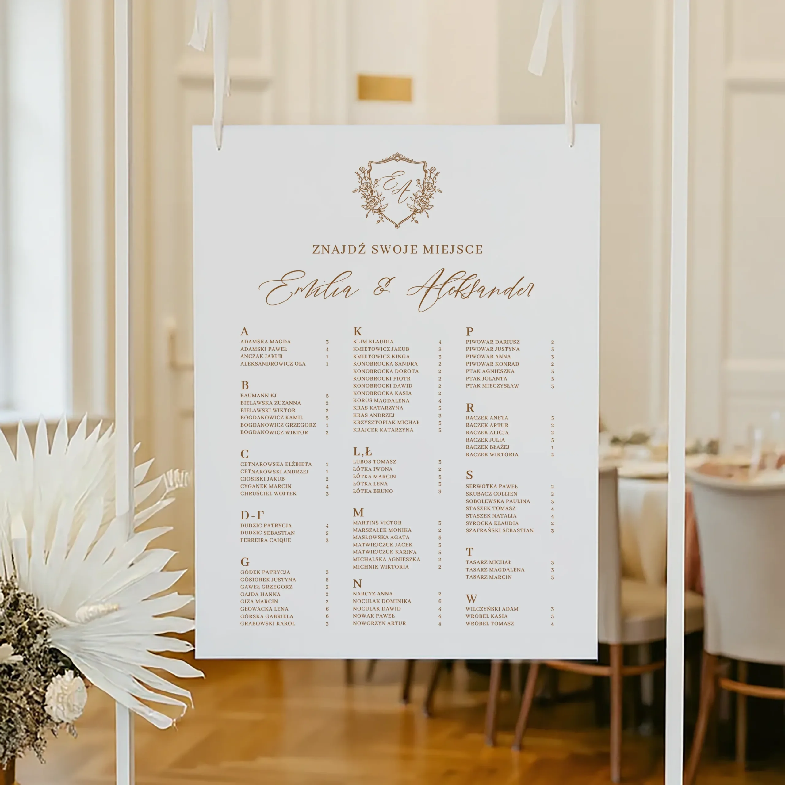 Plan stołów z alfabetycznym układem nazwisk i złotym monogramem, zawieszony na białej ramie.