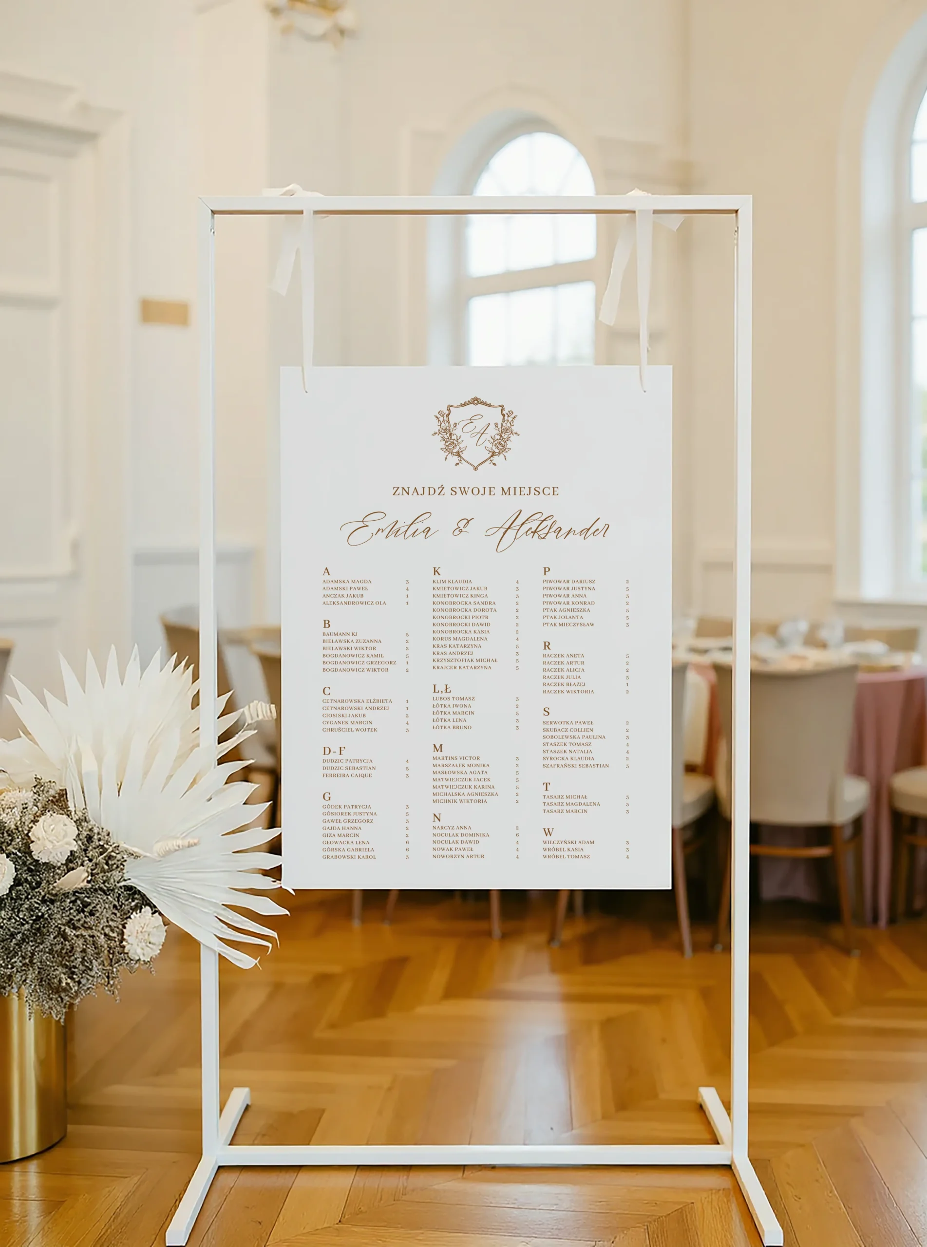 Plan stołów w stylu klasycznym z monogramem i nazwiskami gości ułożonymi alfabetycznie, zawieszony na białym stojaku.