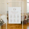 Plan stołów z elegancką czcionką i ozdobnym herbem, zawieszony na stojaku, w otoczeniu klasycznej sali weselnej.