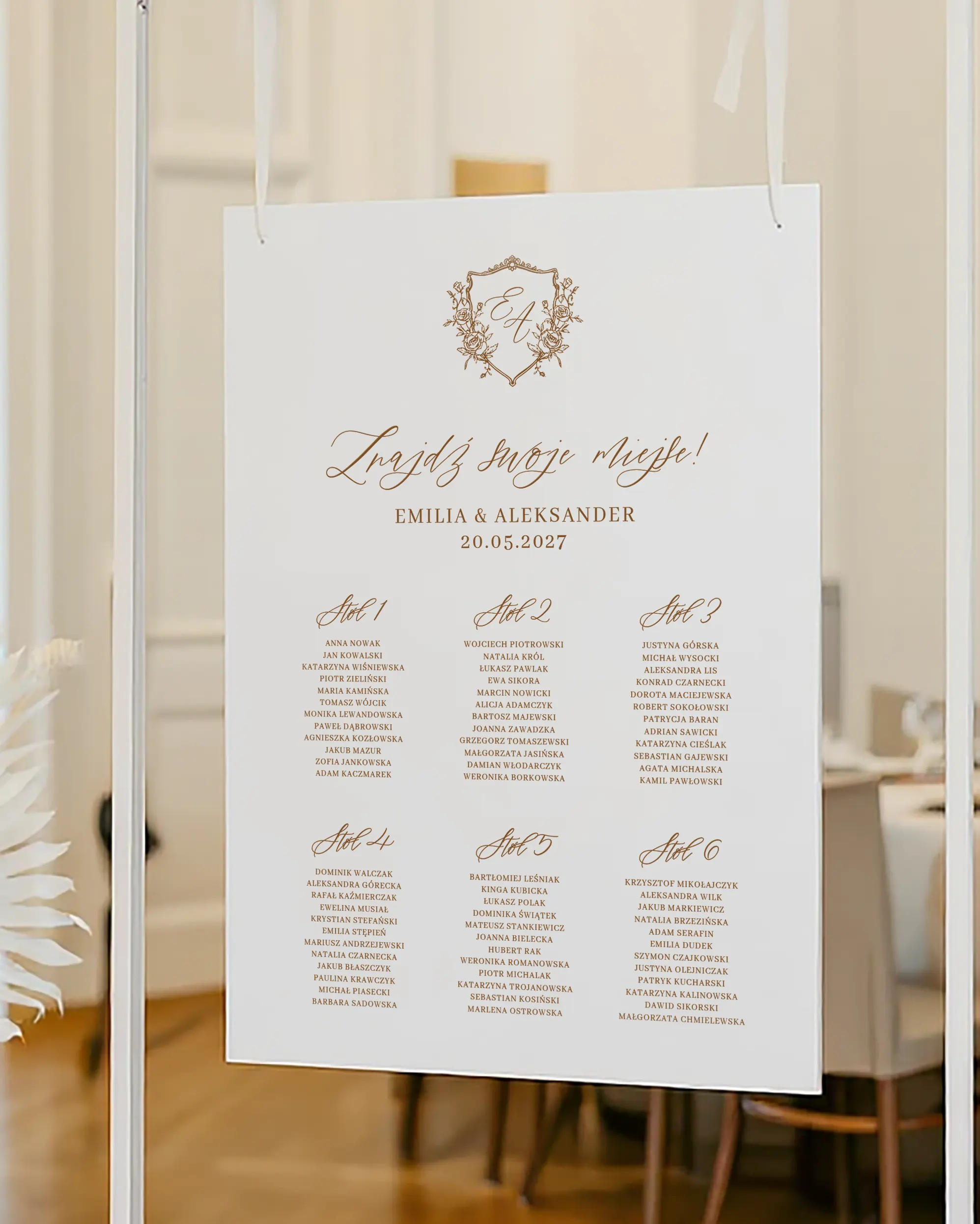 Plan stołów z nazwiskami gości przydzielonymi do konkretnych stołów, zawieszony na eleganckim białym stojaku.