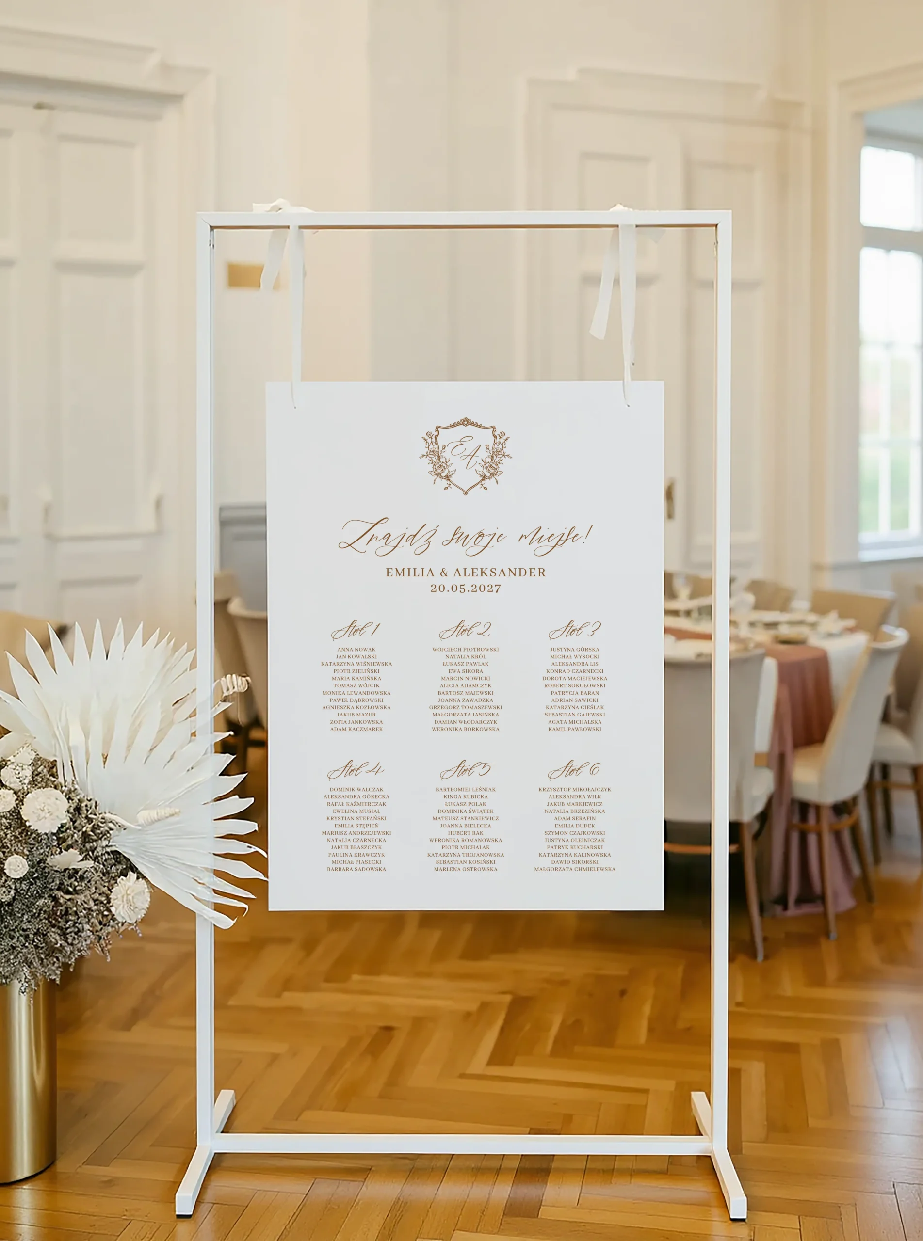 Plan stołów z elegancką czcionką i ozdobnym herbem, zawieszony na stojaku, w otoczeniu klasycznej sali weselnej.