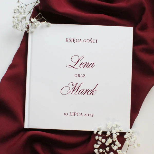 księga gości Burgund w białej oprawie z bordowym nadrukiem imion, na tle bordowego materiału i białych kwiatów.