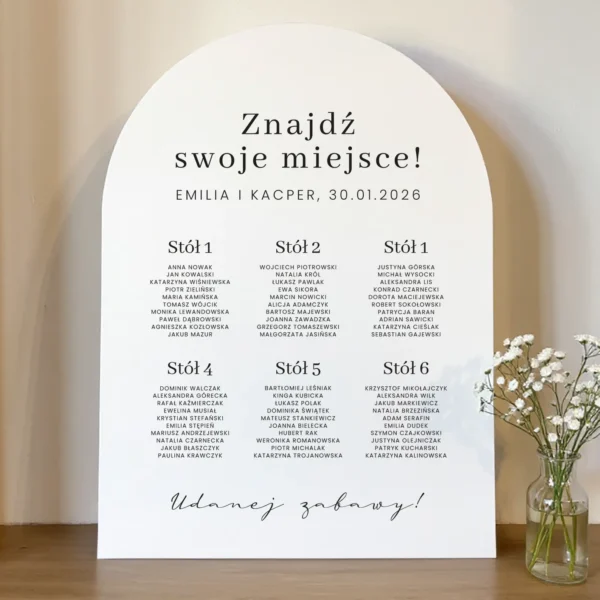 biały plan stołów o zaokrąglonej górze z czarnym napisem „Znajdź swoje miejsce!”, ustawiony na drewnianej półce obok wazonu z gipsówką.