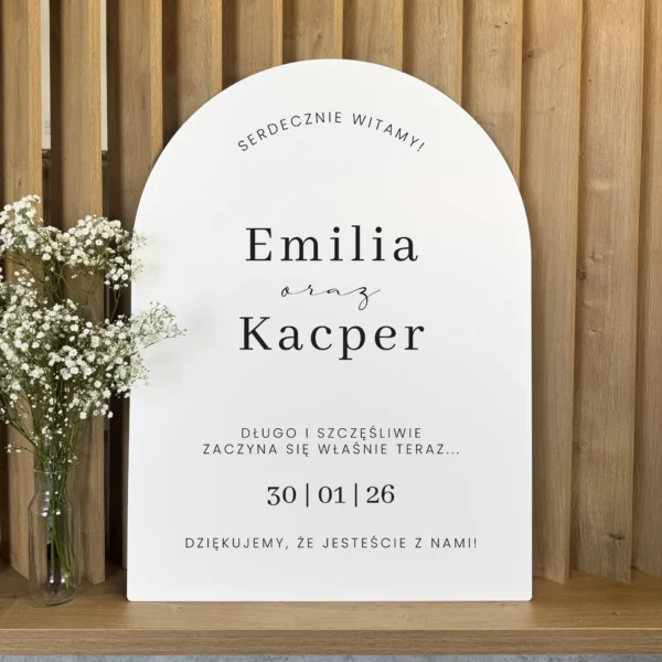 biała tablica powitalna z czarnym napisem i datą, stojąca na tle drewnianej ściany z gipsówką w wazonie.