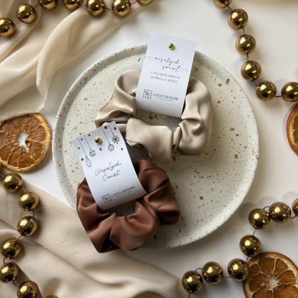 eleganckie gumki scrunchie z zawieszkami świątecznymi z logo firmy, ułożone na talerzyku z ozdobami świątecznymi