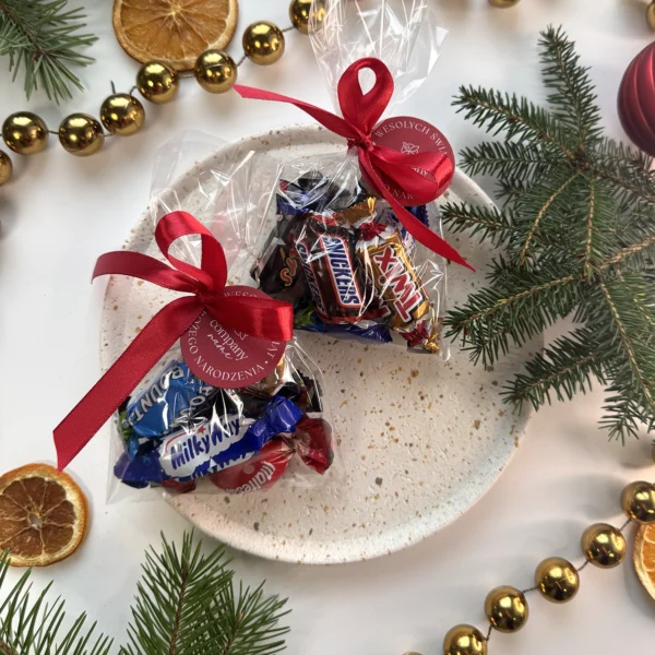 dwa woreczki z cukierkami Celebrations przewiązane czerwoną wstążką z logo firmy na tle świątecznych dekoracji