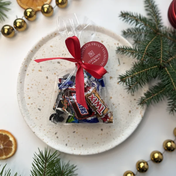 woreczek z cukierkami ozdobiony czerwoną wstążką i zawieszką z napisem wesołych świąt na tle gałązek świerku