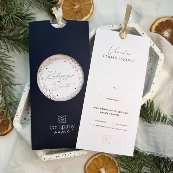 granatowy bon podarunkowy z wysuniętą kartą voucherową na tle gałązek i suszonych pomarańczy