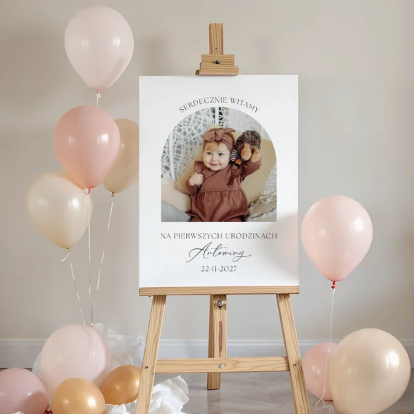 Tablica powitalna na pierwsze urodziny z dużym zdjęciem dziecka, napisem "Serdecznie witamy" oraz pastelowymi balonami w tle.