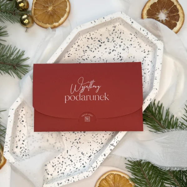 bordowy voucher świąteczny z napisem wyjątkowy podarunek ułożony na białym talerzu w otoczeniu gałązek i suszonych pomarańczy
