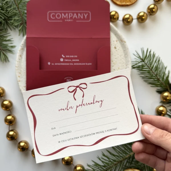 karta podarunkowa z bordową kokardką trzymana w dłoni na tle czerwonego vouchera świątecznego