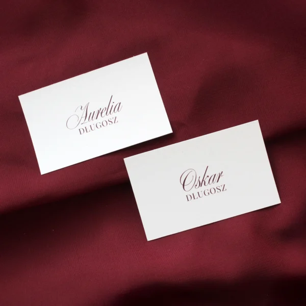 winietki Burgund z imionami gości wydrukowanymi bordowym fontem na białym tle, ułożone na bordowym materiale.