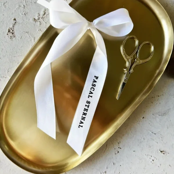 winietka kokardka z imieniem nadrukowanym na białej wstążce, ułożona na złotej tacy obok nożyczek.