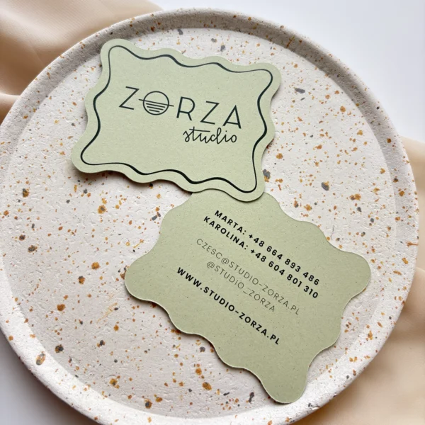 Zestaw wizytówek o falowanym kształcie, z eleganckim logo Zorza Studio, ułożonych na talerzu z efektem terrazzo.