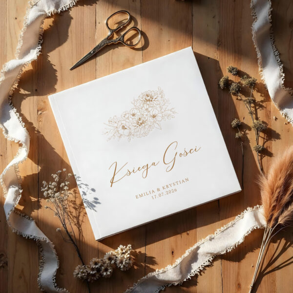 księga gości Dalia w białej oprawie ze złotym napisem i kwiatowym motywem, sfotografowana w ciepłym świetle na drewnianym tle z dekoracjami.