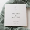 elegancka, biała księga z minimalistycznym monogramem i imionami Pary Młodej, ułożona na delikatnej, zielonej tkaninie