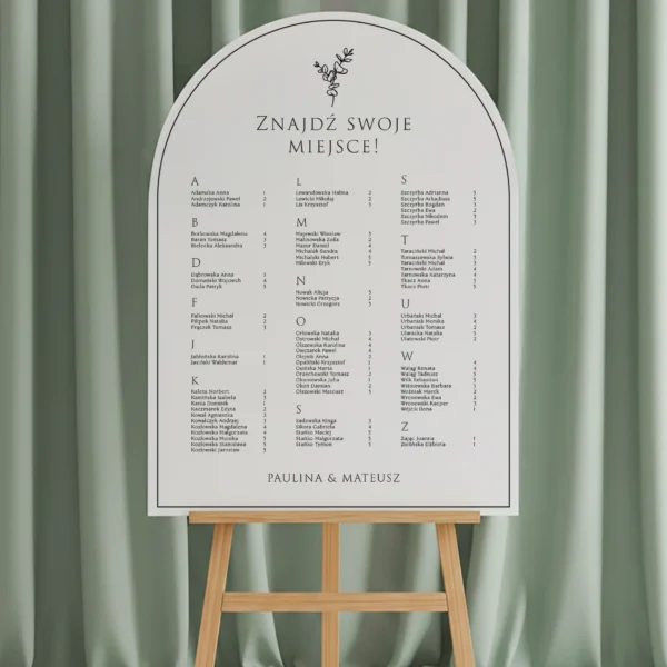 tablica w kształcie łuku z listą gości w układzie alfabetycznym, ustawiona na drewnianej sztaludze na tle zielonej zasłony