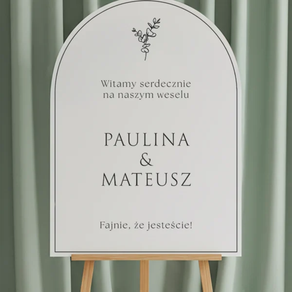 tablica w kształcie łuku z delikatną gałązką eukaliptusa i imionami Pary Młodej, wyeksponowana na sztaludze w eleganckiej aranżacji