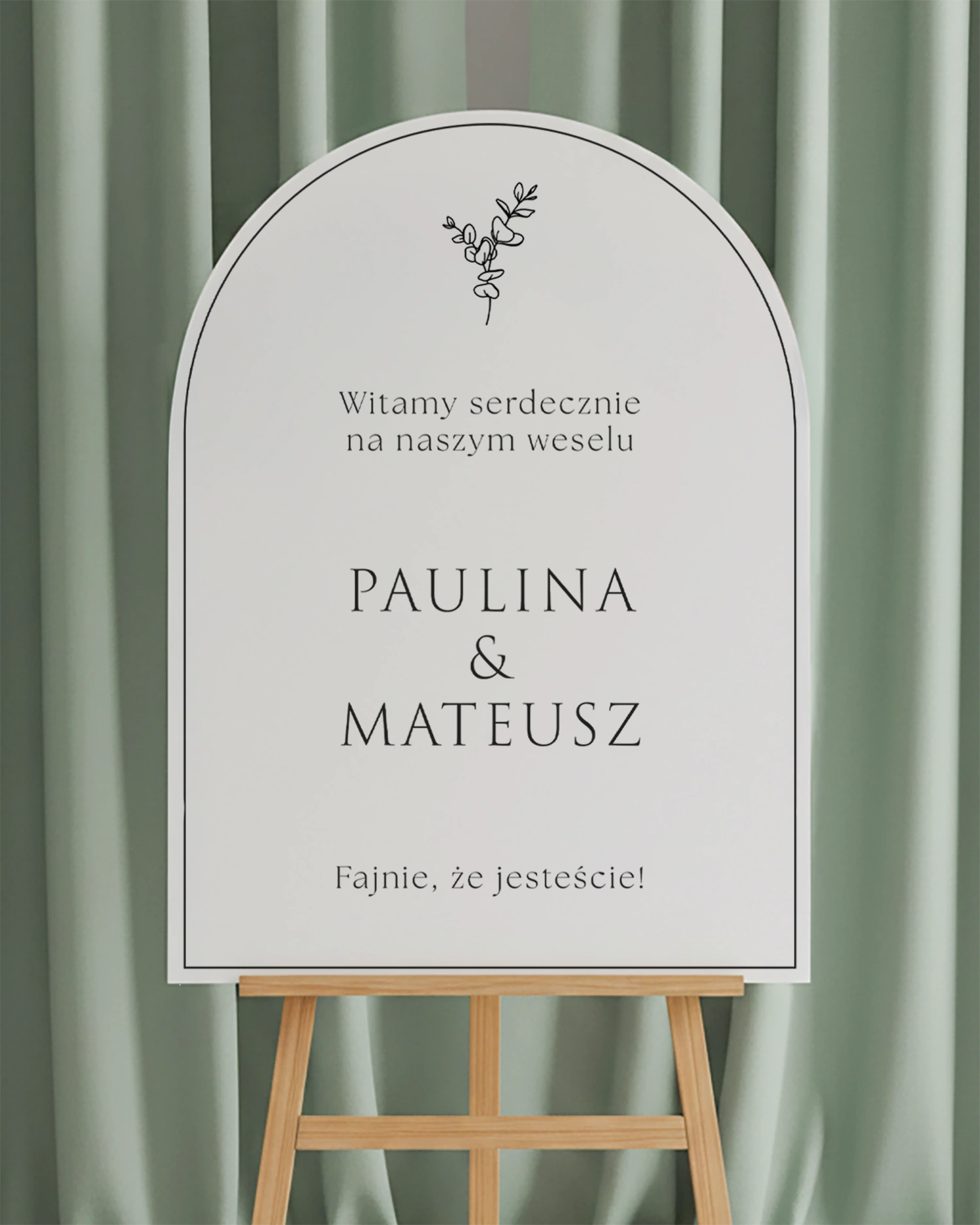 tablica w kształcie łuku z delikatną gałązką eukaliptusa i imionami Pary Młodej, wyeksponowana na sztaludze w eleganckiej aranżacji