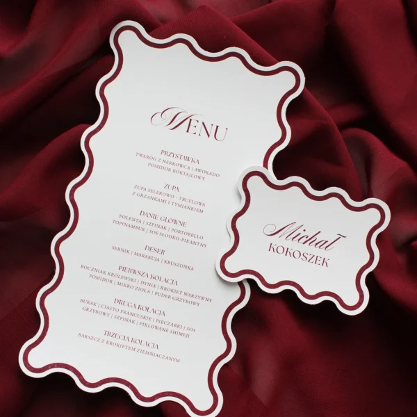 Stylowe menu weselne i winietka z falowanymi brzegami i bordową ramką, na tle bordowej tkaniny.