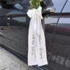 Elegancka dekoracja samochodu ślubnego z białą kokardą, kwiatami i długimi personalizowanymi wstążkami.
