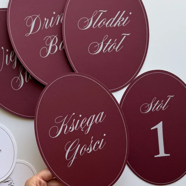 Burgundowe owalne numery stołów weselnych z eleganckimi napisami i delikatną jasną obwódką.