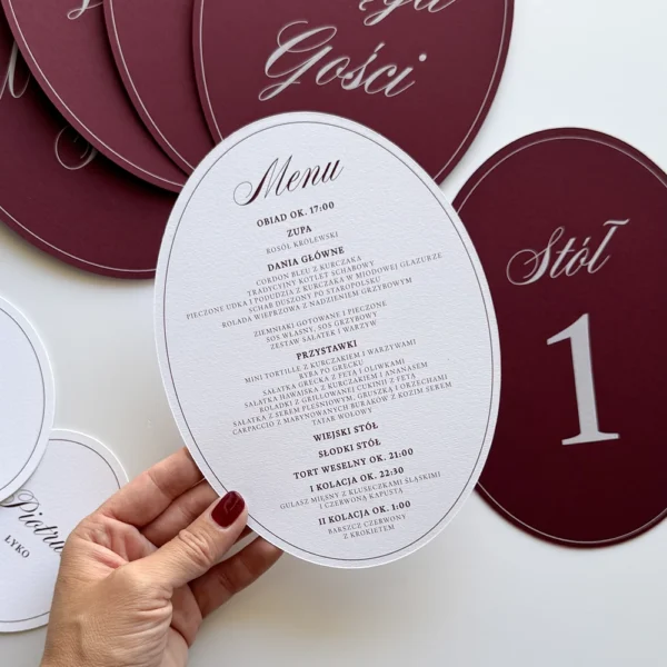 Owalne menu weselne w śmietankowym kolorze z burgundowym nadrukiem, na tle eleganckiej papeterii ślubnej.