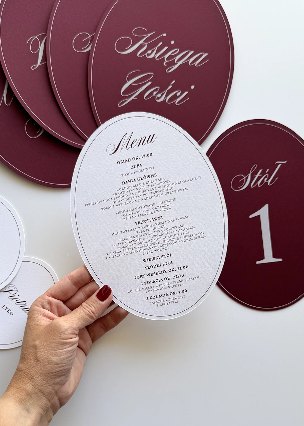 Owalne menu weselne w śmietankowym kolorze z burgundowym nadrukiem, na tle eleganckiej papeterii ślubnej.